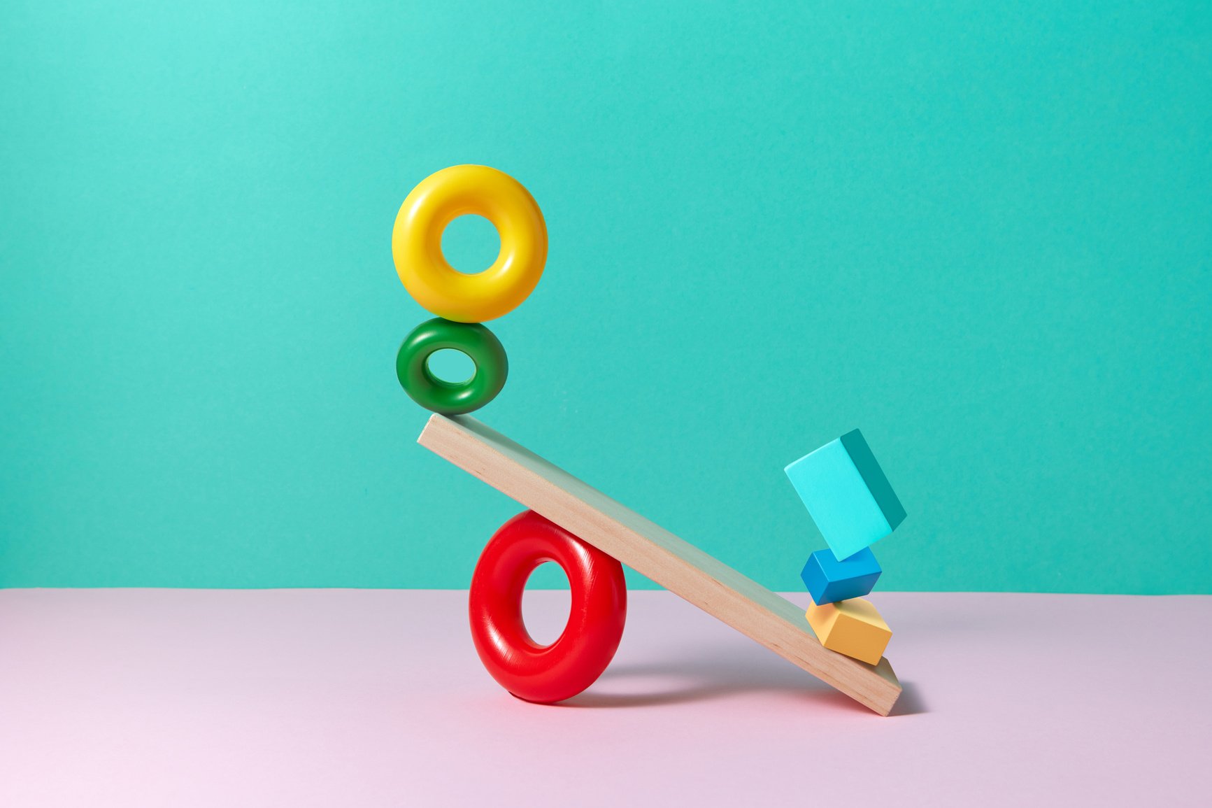 Colourful 3D rings and cubes balance on a wooden plank like a seesaw, with the right-hand side weighing far heavier than the left